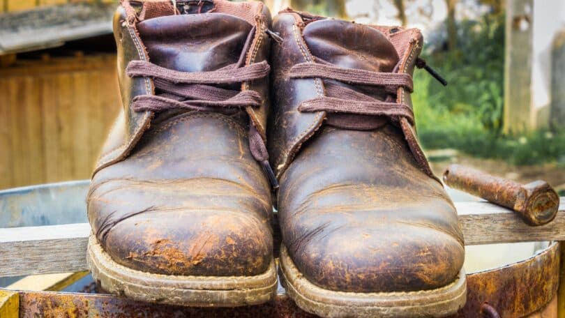 Imagem de um sapato velho, uma bota marrom, sobre um latão de ferro. O sapato está com marcas e sujo de barro.