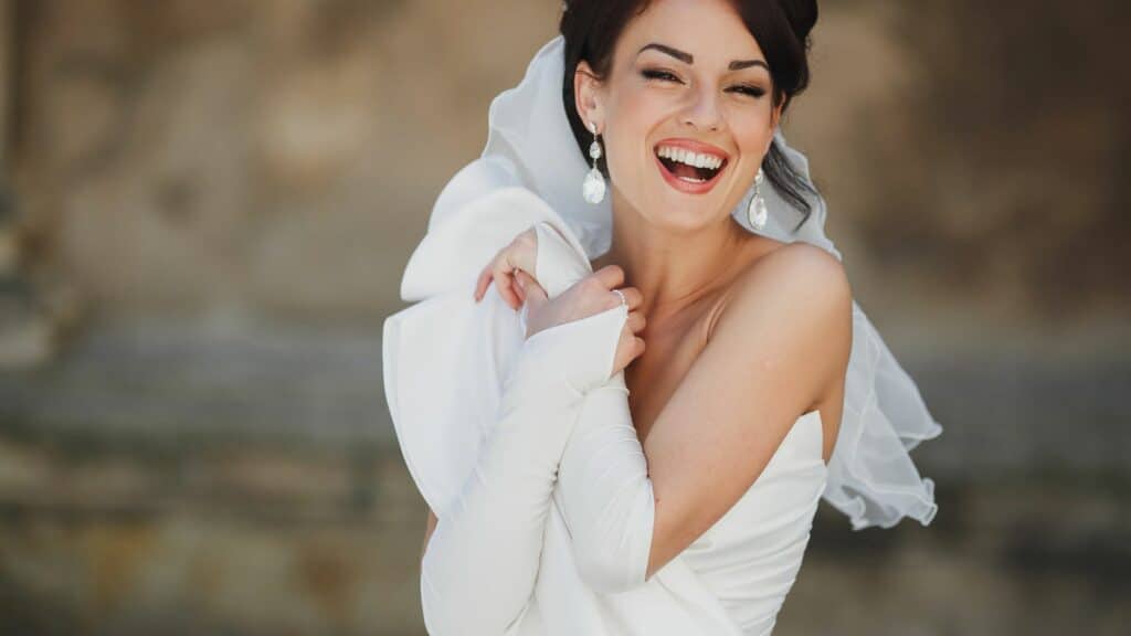Imagem de uma noiva muito feliz, sorridente e radiante. Ela usa um lindo vestido branco, simples, porém elegante e um belíssimo par de brincos em formato de gota.