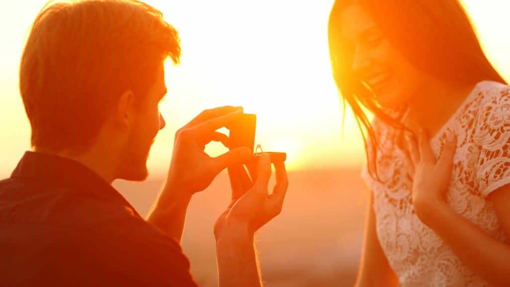 Imagem de um lindo pôr do Sol ao fundo e em destaque um homem, abrindo uma caixa de anel e fazendo o pedido de casamento para a sua noiva.