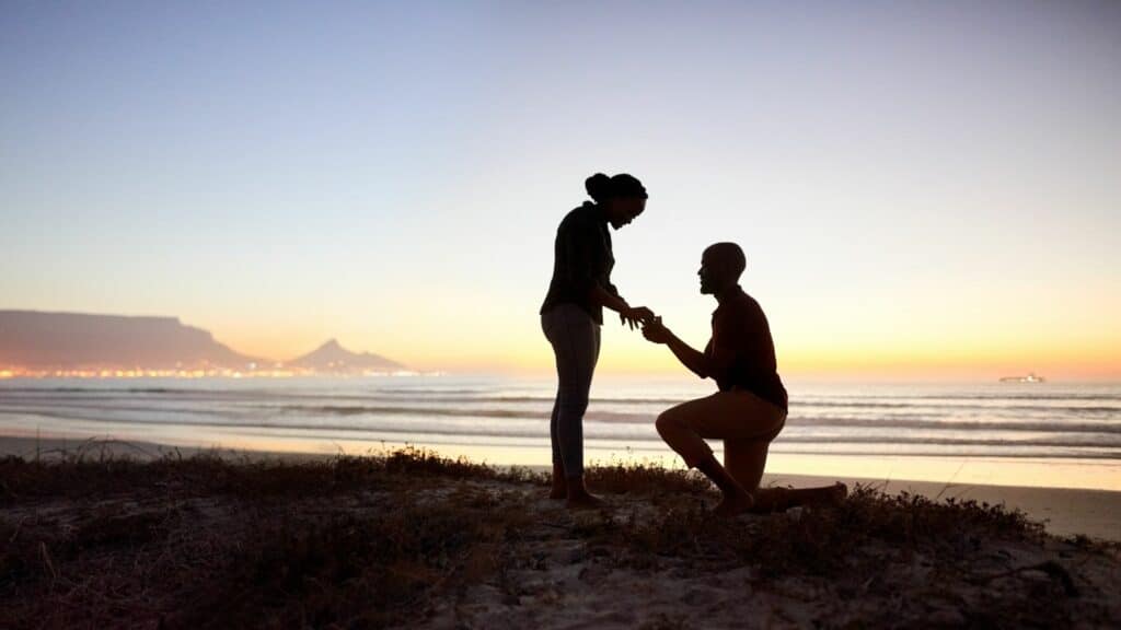 Imagem de uma praia ao fundo e em destaque um homem e uma mulher. O homem está ajoelhado, fazendo um pedido de casamento para a sua noiva.