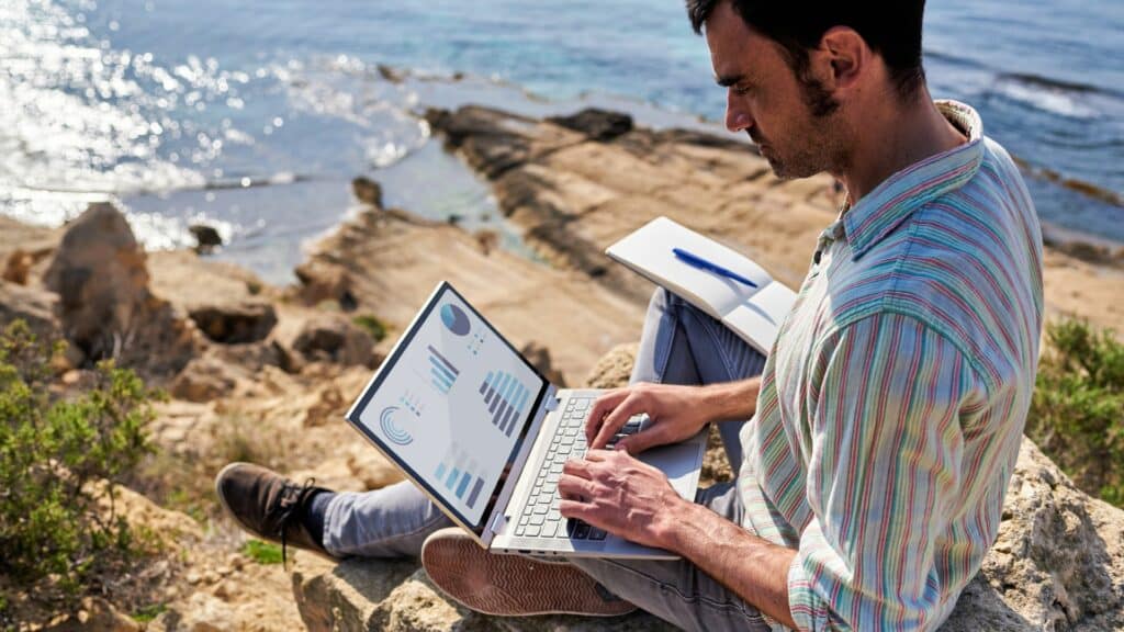 Imagem de um homem sentada sobre uma pedra em uma montanha em frente a um lago. Ele está trabalhando em seu notebook de forma remota.