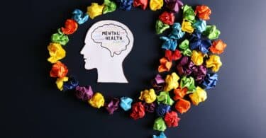 Imagem do contorno de uma cabeça humano onde está escrito Saúde Mental em inglês (Mental Health). Em volta do contorno, vários papéis coloridos amassados.