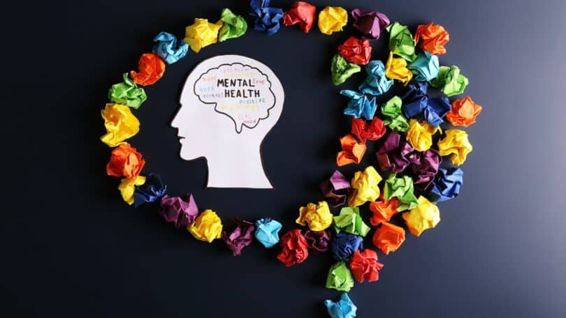 Imagem do contorno de uma cabeça humano onde está escrito Saúde Mental em inglês (Mental Health). Em volta do contorno, vários papéis coloridos amassados.