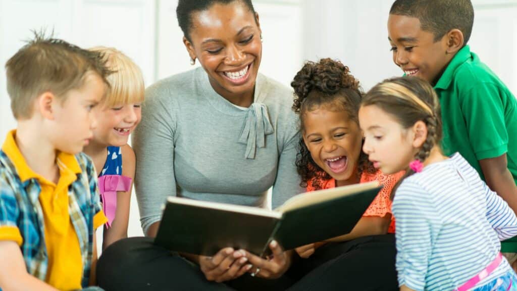 Imagem de uma mulher preta com um livro de histórias nas mãos, lendo contos para as crianças que estão em volta dela.