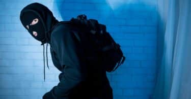 Imagem de um assaltante usando roupas pretas e uma máscara com capuz. Ele leva uma mochila preta nas costas.