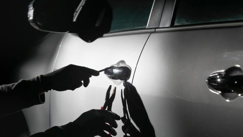 Imagem em preto e branco e em destaque as mãos de um assaltante tentando abrir a porta de um carro.