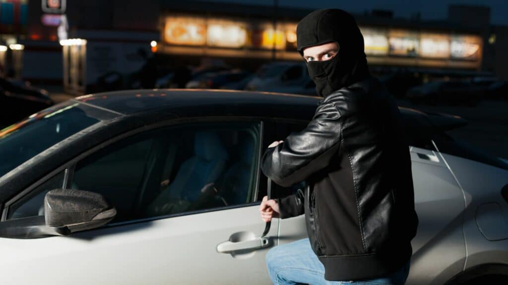 Imagem de ladrão de carros, usando calça jeans, jaqueta de couro e um gorro na cor preta. Ele está com uma faca na mão, tentando abrir a porta do carro.