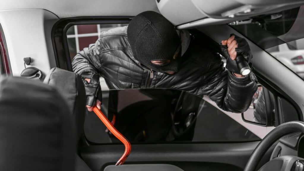 Imagem de um ladrão entrando pela janela do carro para fazer o furto do mesmo. Ele segura uma lanterna e um pé de cabras.
