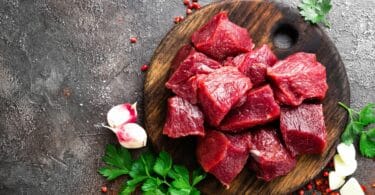 Imagem de uma tábua de madeira e sobre ela, vários pedaços de carne crua vermelha. Ao lado, temperos como salsinha, alho e pimenta rosa.