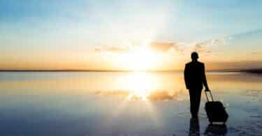 Imagem de um lindo rio e ao fundo, o pôr do sol. Em destaque, um homem puxando a sua mala. A foto traz o conceito de que a vida é uma viagem.