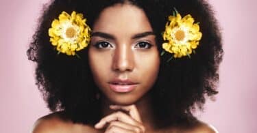Imagem de uma linda mulher preta com seus cabelos naturais decorados com duas flores amarelas, uma de cada lado. A foto traz o conceito de autenticidade, de ser autêntico.