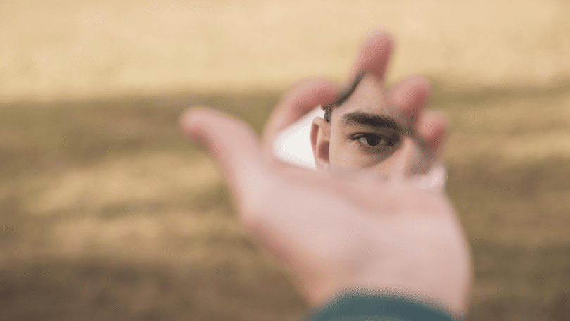 O olhar de um homem está sendo refletido em um pedaço de espelho quebrado, o qual ele está segurando.
