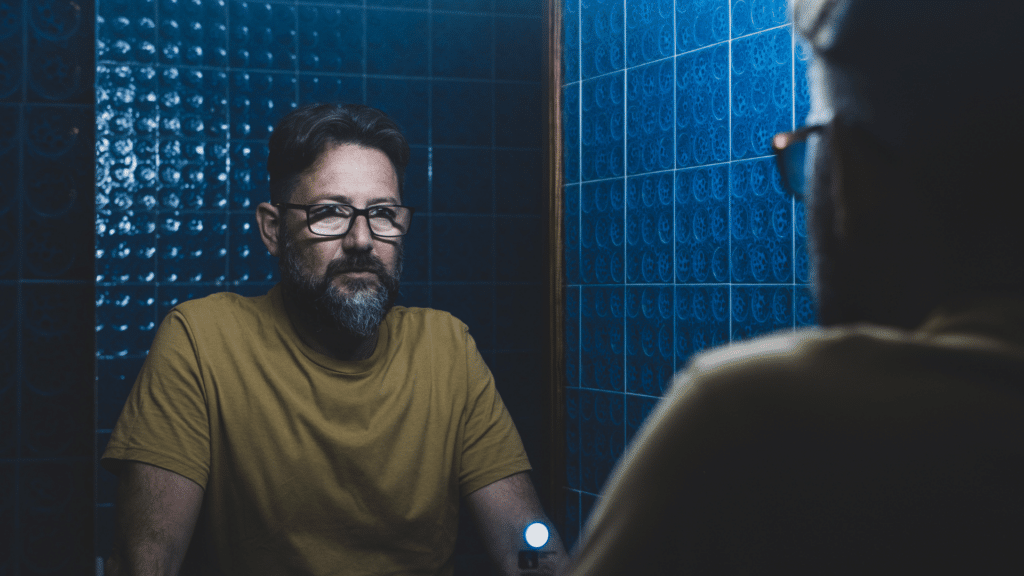 Um homem que usa óculos e tem barba se olha no espelho e encara o seu reflexo.