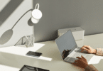 As mãos de uma mulher digitando em um notebook estão em destaque na imagem. Há uma uma mesa minimalista com poucos objetos, a maioria na cor preta e branca.