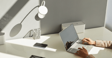 As mãos de uma mulher digitando em um notebook estão em destaque na imagem. Há uma uma mesa minimalista com poucos objetos, a maioria na cor preta e branca.