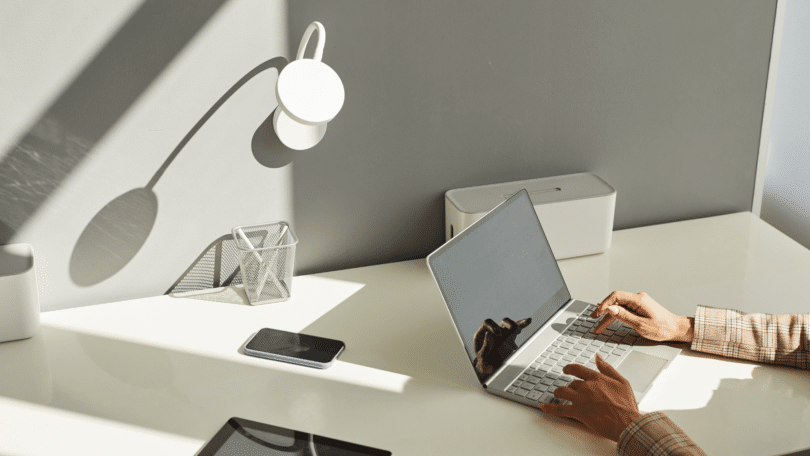 As mãos de uma mulher digitando em um notebook estão em destaque na imagem. Há uma uma mesa minimalista com poucos objetos, a maioria na cor preta e branca.