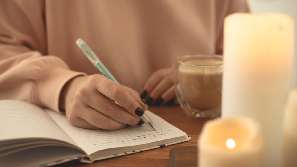 A mão de uma mulher escrevendo em seu caderno está em destaque na imagem. Há velas ao lado.