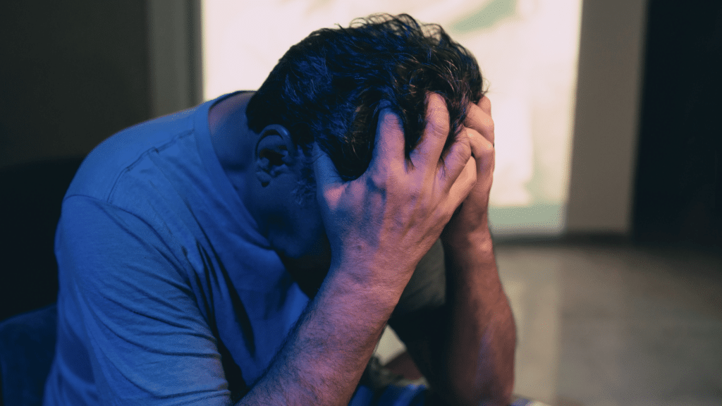 Um homem está cabisbaixo e com as duas mãos na cabeça, indicando estar triste.
