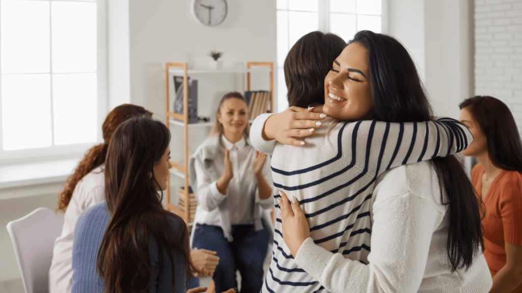 Duas mulheres estão se abraçando no foco da imagem, indicando apoio. Atrás delas, há um grupo com outras mulheres sentadas.