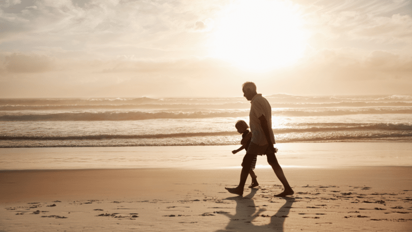 Um avô e seu neto estão caminhando na praia sob a luz do pôr do sol.