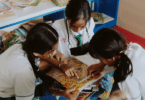 Três alunas pequenas estão lendo um livro juntas na sala de aula.
