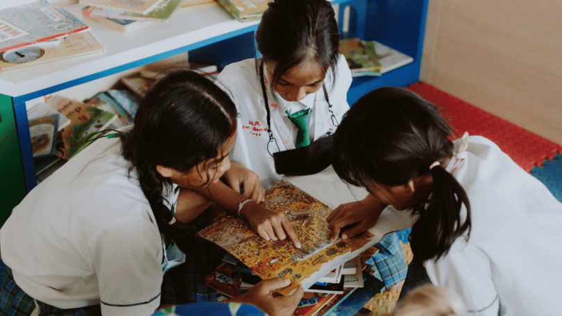 Três alunas pequenas estão lendo um livro juntas na sala de aula.