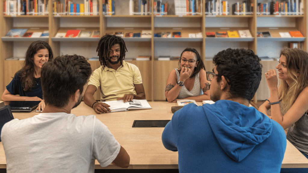 Seis jovens universitários estão sentados e debatendo um assunto em uma biblioteca.