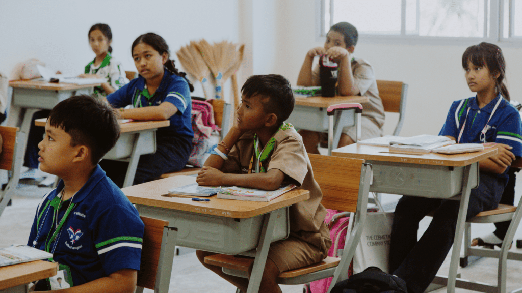 Seis crianças estão sentadas em suas cadeiras em uma sala de aula. Elas prestam atenção no que a professora fala.