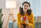 Uma mulher está sentada no seu sofá com uma xícara na mão. Ela escuta música nos seus fones de ouvido, fecha os olhos e sente a música.