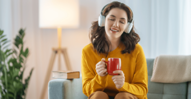 Uma mulher está sentada no seu sofá com uma xícara na mão. Ela escuta música nos seus fones de ouvido, fecha os olhos e sente a música.
