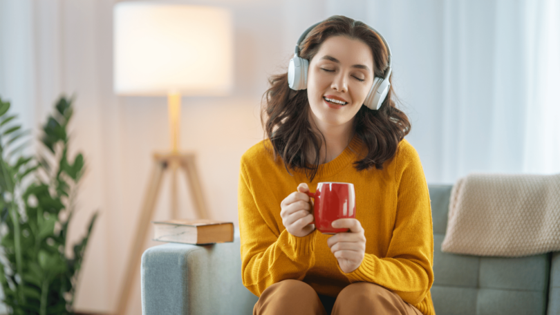 Uma mulher está sentada no seu sofá com uma xícara na mão. Ela escuta música nos seus fones de ouvido, fecha os olhos e sente a música.