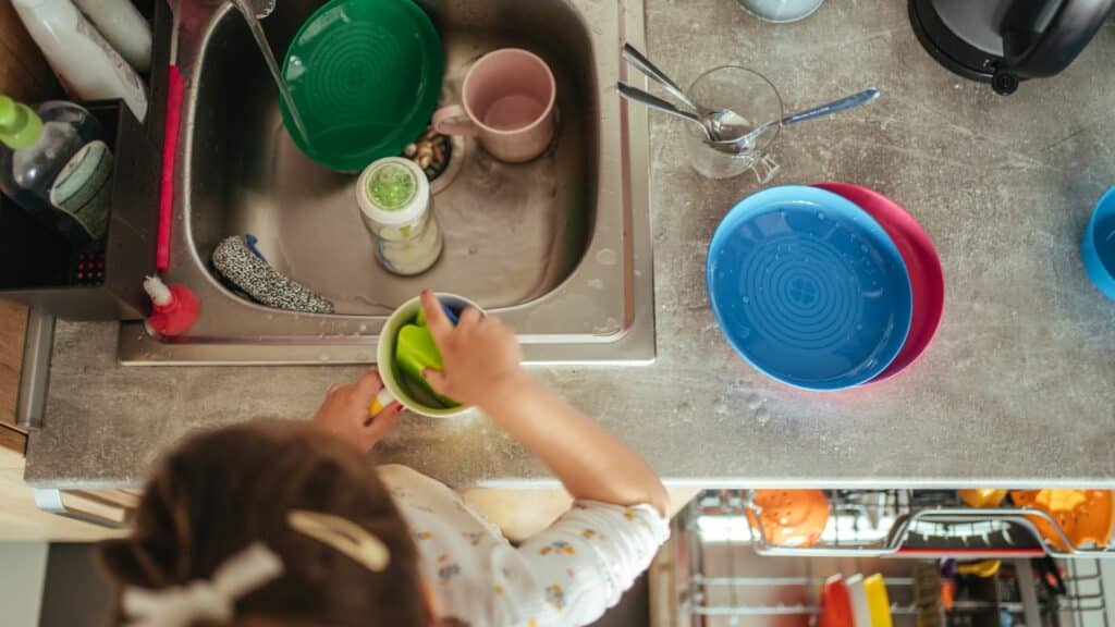 Imagem de uma criança na pia de uma cozinha, lavando várias louças.
