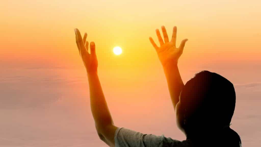 Imagem de fundo com um lindo Sol e em destaque uma pessoa olhando para ele com as mãos ao alto.
