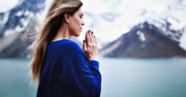 Imagem de uma mulher de frente para uma montanha coberta com gelo. Ela está com as mãos em posição de fé e com os olhos fechados.