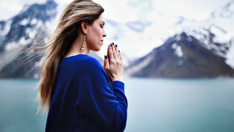 Imagem de uma mulher de frente para uma montanha coberta com gelo. Ela está com as mãos em posição de fé e com os olhos fechados.