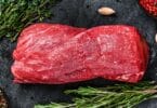 Imagem de uma linda peça de carne de boi. Ao lado, ramos de alecrim, orégano, pimenta rosa e preta e alguns dentes de alho.