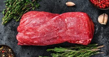 Imagem de uma linda peça de carne de boi. Ao lado, ramos de alecrim, orégano, pimenta rosa e preta e alguns dentes de alho.