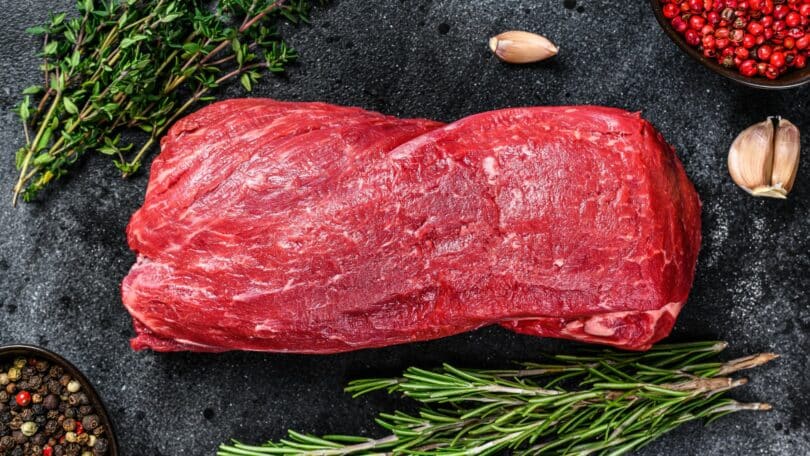 Imagem de uma linda peça de carne de boi. Ao lado, ramos de alecrim, orégano, pimenta rosa e preta e alguns dentes de alho.