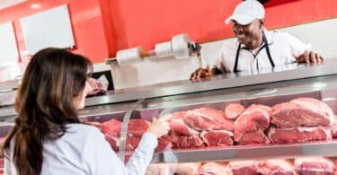 Imagem de uma mulher comprando carne de boi em um açougue. Ela está sendo atendida por um açougueiro.
