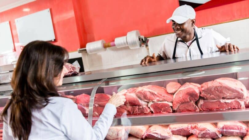 Imagem de uma mulher comprando carne de boi em um açougue. Ela está sendo atendida por um açougueiro.