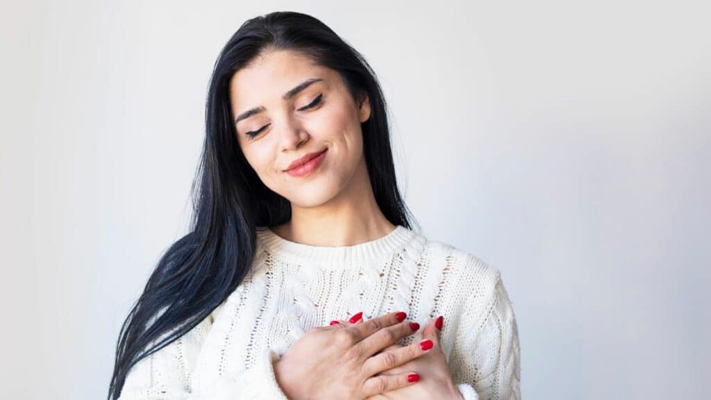 Imagem de uma mulher de olhos fechados e sorrindo levemente. Ela está com as duas mãos sobre o seu coração. A foto traz o conceito de autoconhecimento e autocuidado.