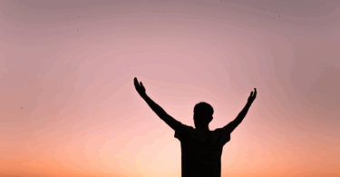 Um homem está com os dois braços abertos e levantados em um ambiente de ar livre, indicando gratidão. Ao fundo, há o céu alaranjado do pôr do sol.