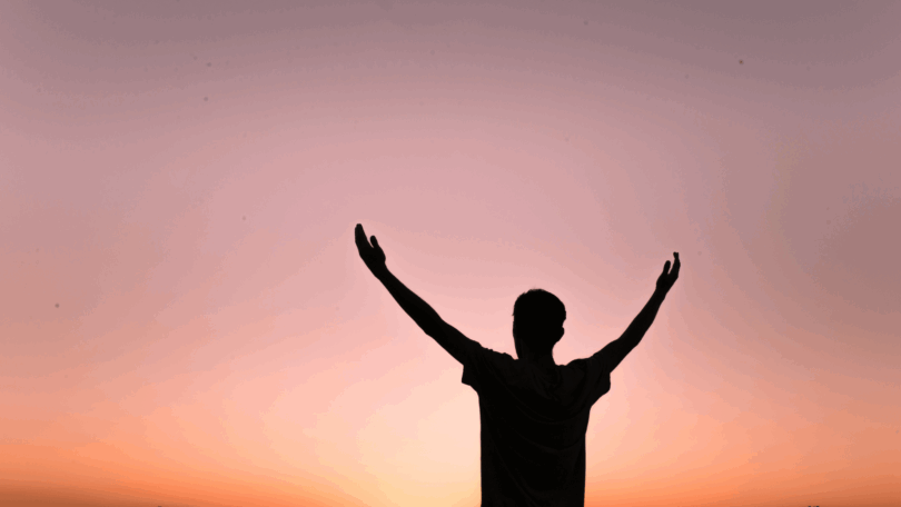 Um homem está com os dois braços abertos e levantados em um ambiente de ar livre, indicando gratidão. Ao fundo, há o céu alaranjado do pôr do sol.