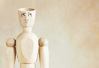 Um boneco de madeira com um rosto desenhado está no foco da imagem. O boneco olha para cima, pensativo ou em dúvida. A parte de cima da cabeça dele está cortada, indicando ser um boneco ou homem de cabeça vazia.