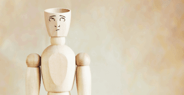 Um boneco de madeira com um rosto desenhado está no foco da imagem. O boneco olha para cima, pensativo ou em dúvida. A parte de cima da cabeça dele está cortada, indicando ser um boneco ou homem de cabeça vazia.