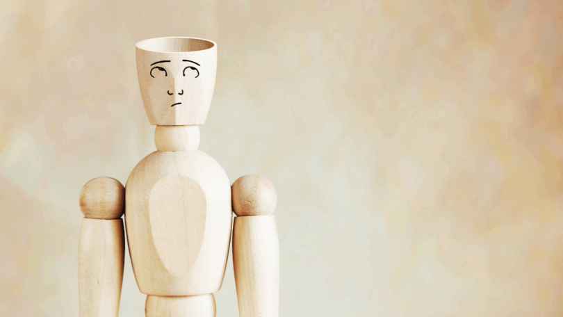 Um boneco de madeira com um rosto desenhado está no foco da imagem. O boneco olha para cima, pensativo ou em dúvida. A parte de cima da cabeça dele está cortada, indicando ser um boneco ou homem de cabeça vazia.