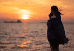 Uma mulher jovem, triste e reflexiva está de frente para o mar. Ela está enrolada em um cobertor, o vento bate no seu cabelo e ela observa o pôr do sol.