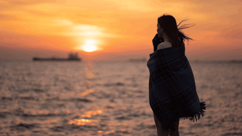 Uma mulher jovem, triste e reflexiva está de frente para o mar. Ela está enrolada em um cobertor, o vento bate no seu cabelo e ela observa o pôr do sol.