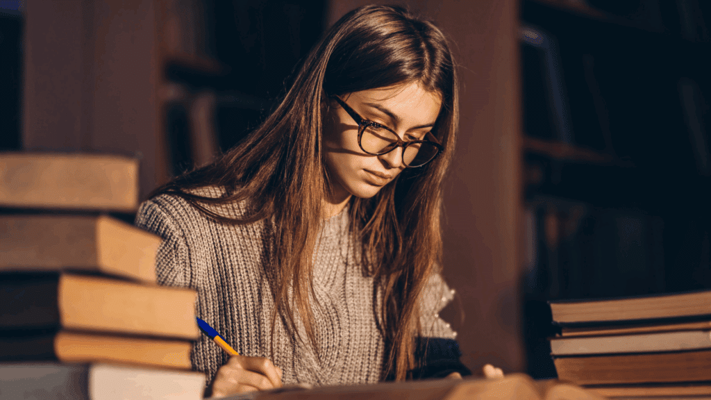 Uma mulher jovem está estudando em uma biblioteca. Ela segura a caneta e lê um livro.