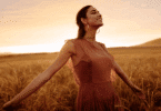 Uma mulher jovem está em um campo durante o pôr do sol. Ela levanta o rosto e sorri sentindo a brisa do vento. Ela está com os dois braços abertos.